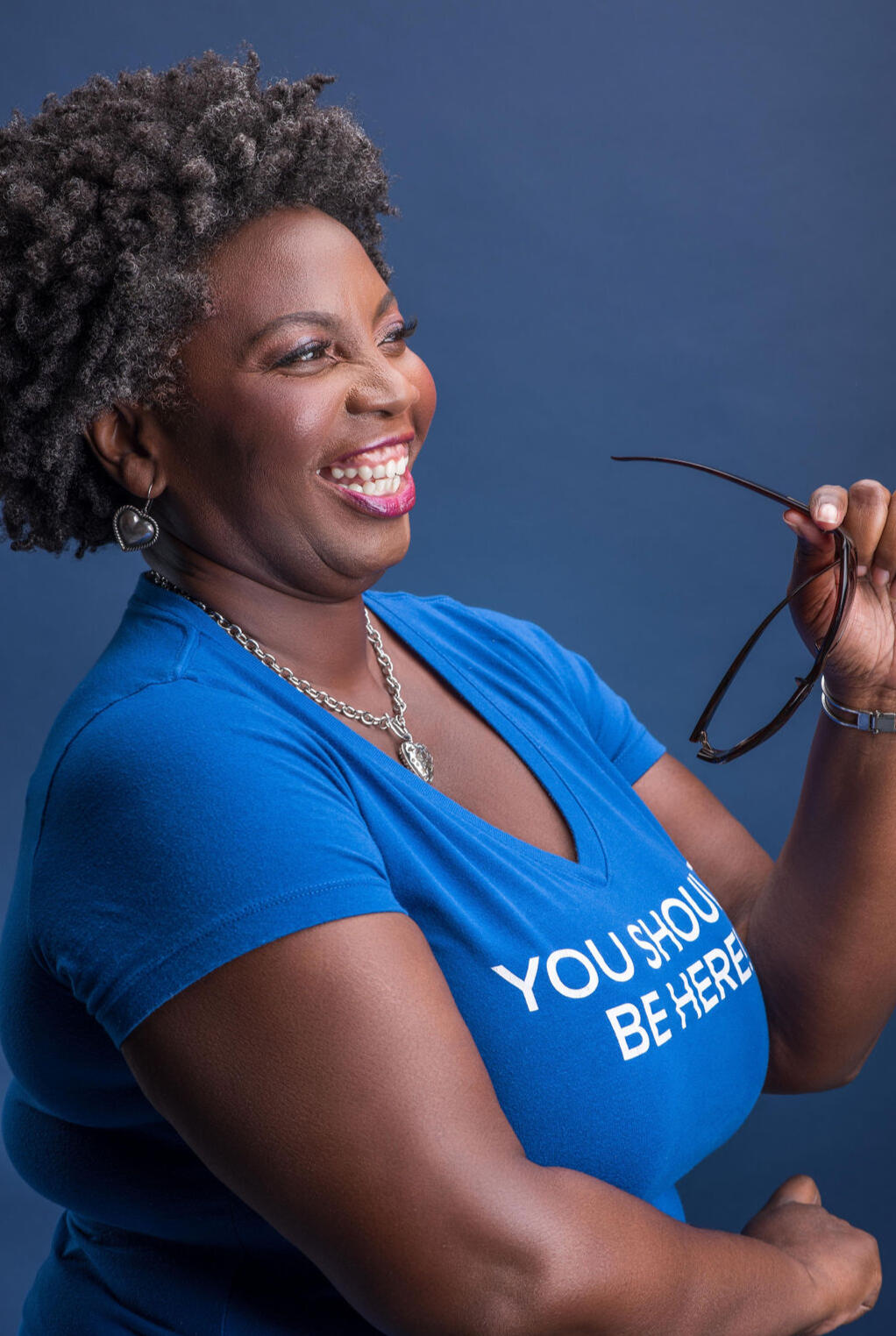 Regina Nunnally Regina wearing a blue t-shirt with the words "you should be hear" printed in white, Regina is holding sunglasses in one hand and has a happy smile.