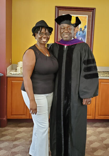 Regina Nunnally with Attorney Benjamin Crump. Regina is wearing a black hat and black shirt with White pants, Bejamin is standing next to her wearing a graduation gown in a room at Bethune Cookman University