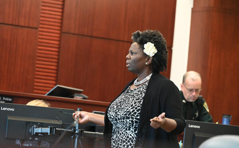 Regina in Courtroom Regina, as Public Defender, is speaking in a courtroom with court clerks behind her, sitting at desks with monitors around them. Regina is wearing a black and white top with a black cardigan,