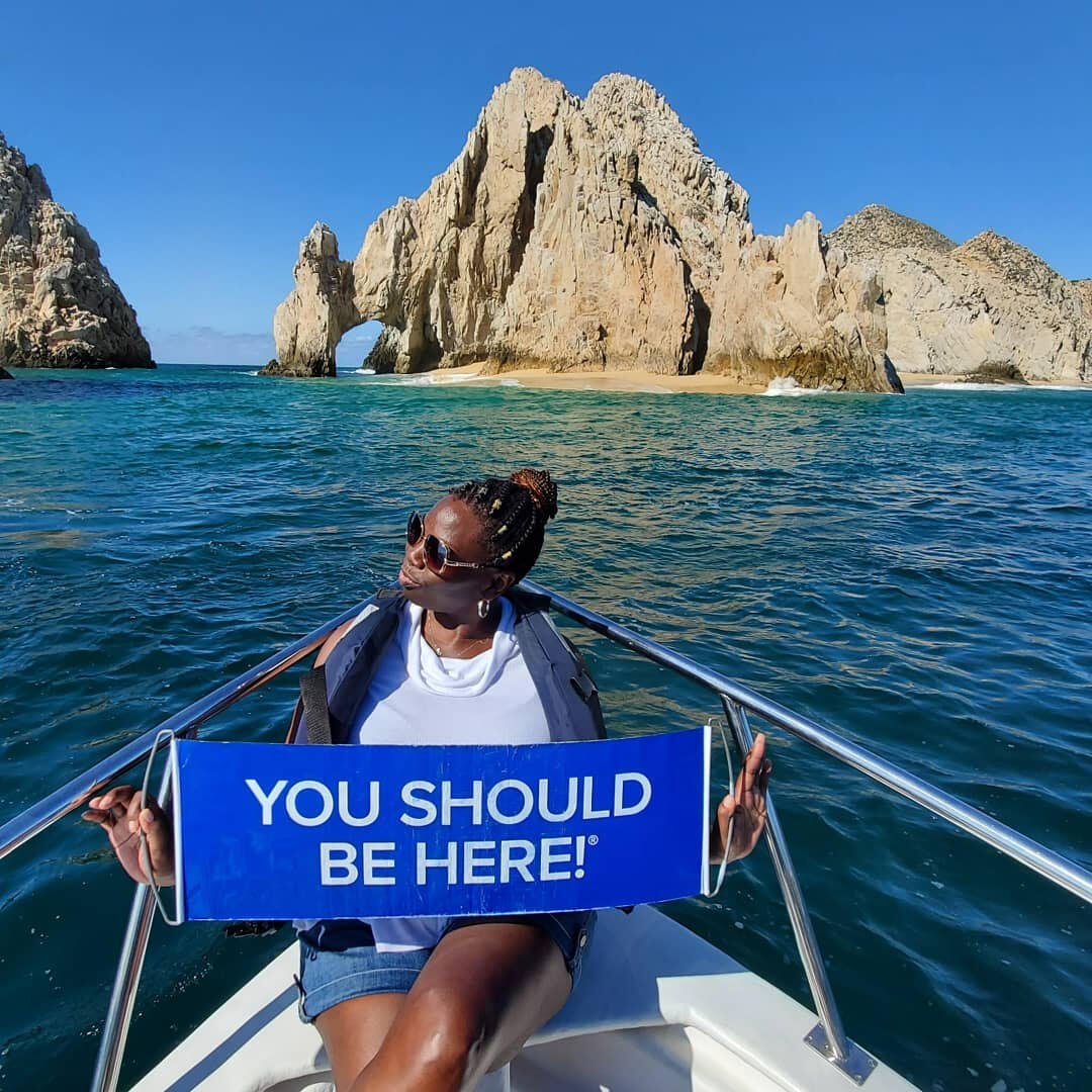 Regina sat in a small boat on the ocean holding a banner saying "You should be here"